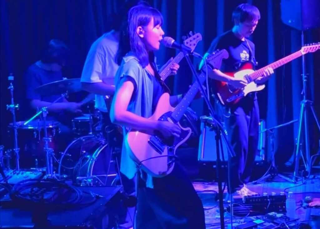 A performer performing live on stage with a local band in Bangkok, playing electric guitar and singing under blue stage lights at an intimate music venue