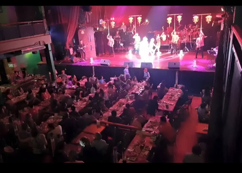 Crowded dinner theater with live band and traditional dance performance on stage.