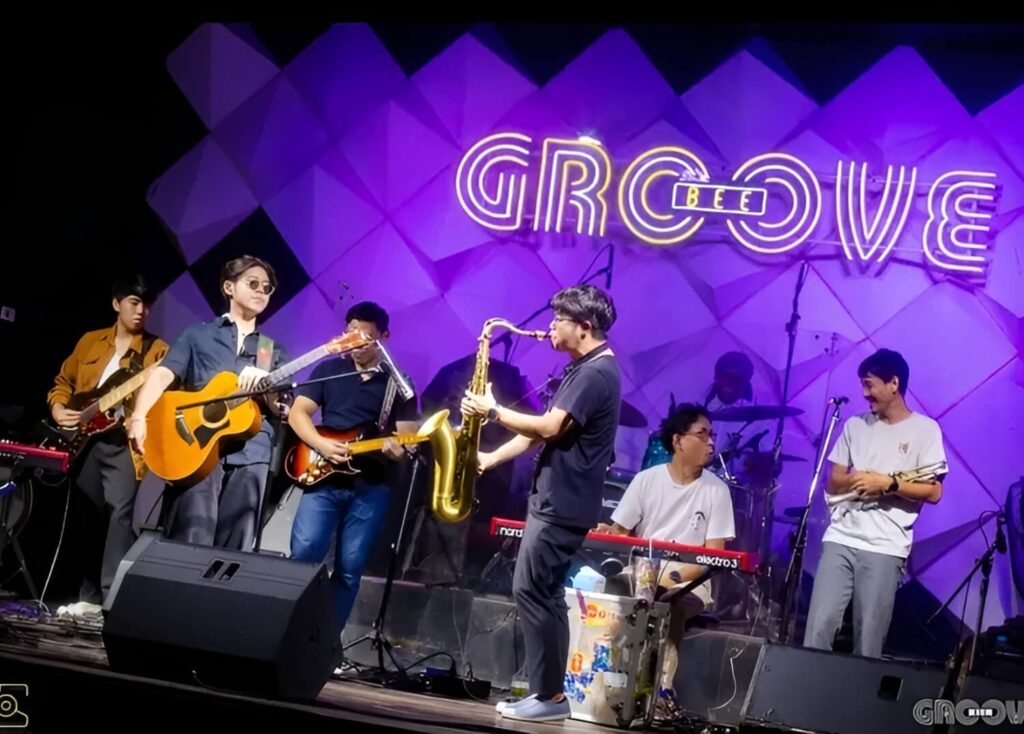 Live band performing on stage under purple lights with 'GROOVE' neon sign in the background.