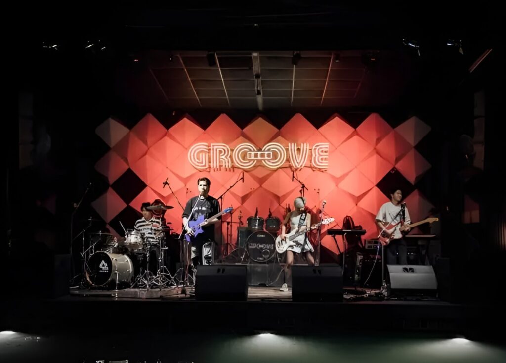 Rock band performing under red lighting at GROOVE stage with vibrant geometric backdrop.
